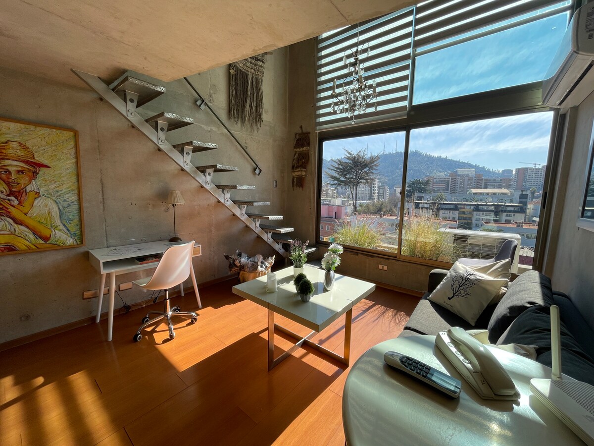 A bright living space features a staircase leading to an upper level. Large windows provide natural light and offer a view of the city skyline. A simple desk and chair, along with a coffee table and a sofa, create a functional area for relaxation or work.