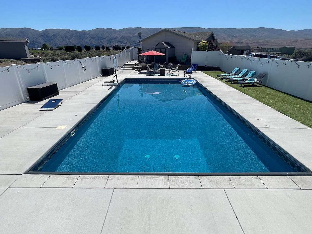 A large heated swimming pool is surrounded by a spacious patio area with sun loungers and an umbrella for shade. The clear blue water reflects the sunny sky, while a well-maintained grass area is visible nearby, providing space for leisure activities.