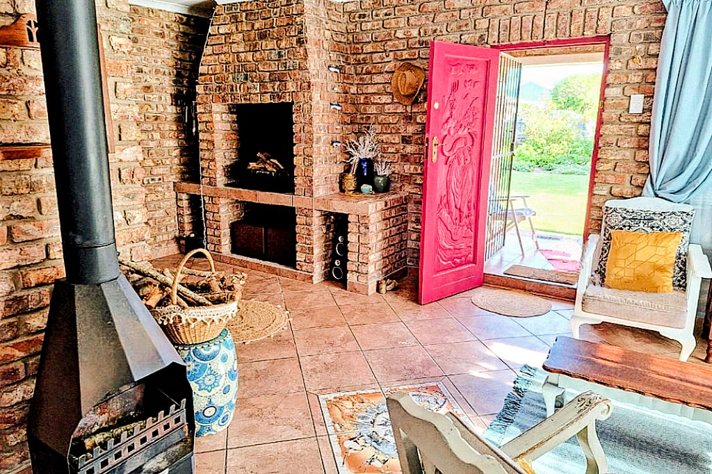 A warm brick interior showcases a well-defined living space with a fireplace and built-in braai. The red front door opens to a lush garden, while a basket of firewood is positioned nearby, complementing the rustic decor. Sunlight streams in, highlighting the inviting atmosphere.