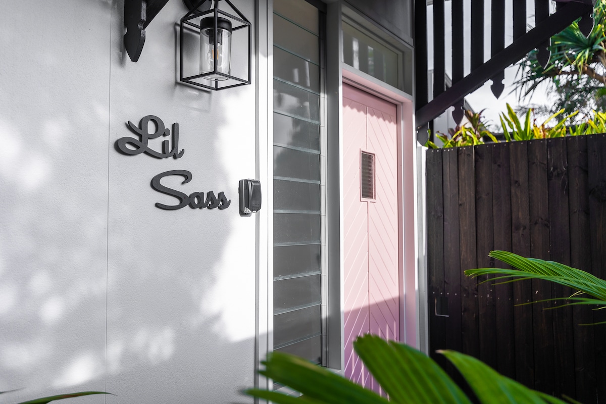 The entrance to Lil Sass is highlighted by a pink door, surrounded by lush greenery and framed by a stylish light fixture. The sign designating the property adds a charming touch, while the surrounding wall offers privacy and a welcoming feel.