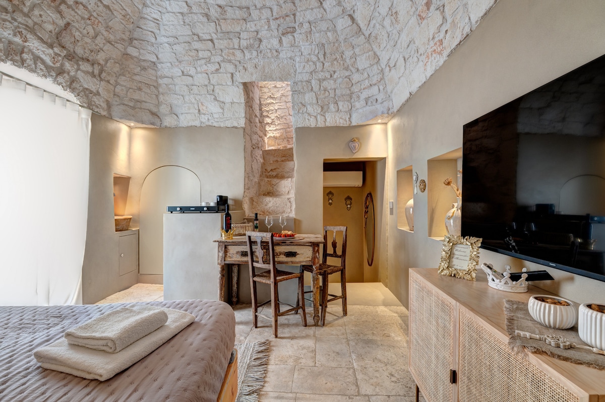 The interior space features a unique stone vault ceiling and soft ambient lighting. A cozy sleeping area with a spacious bed is visible, accompanied by an elegant wooden table with chairs. A smart TV and modern kitchen area are seamlessly integrated into the design.
