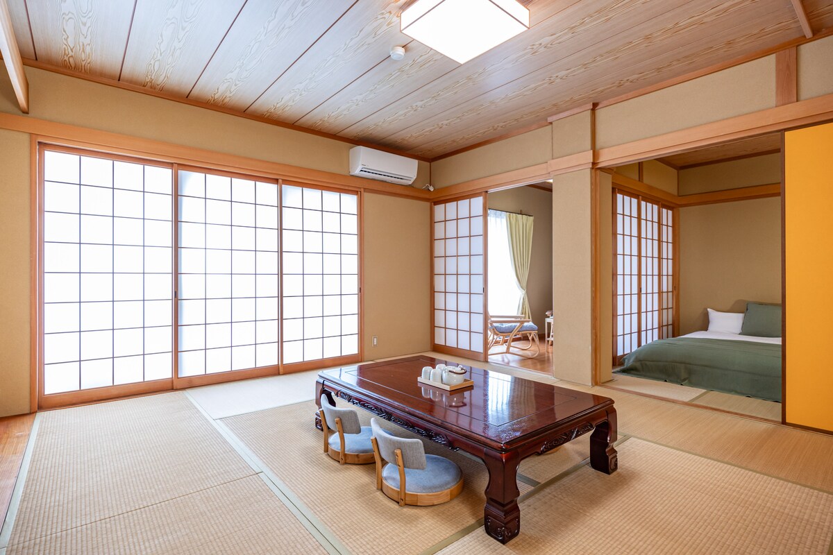 A spacious living area features traditional tatami flooring and large shoji screens, allowing natural light to fill the space. A low wooden table is surrounded by cushioned seating, while a sleeping area with a double bed is visible through an adjoining room.