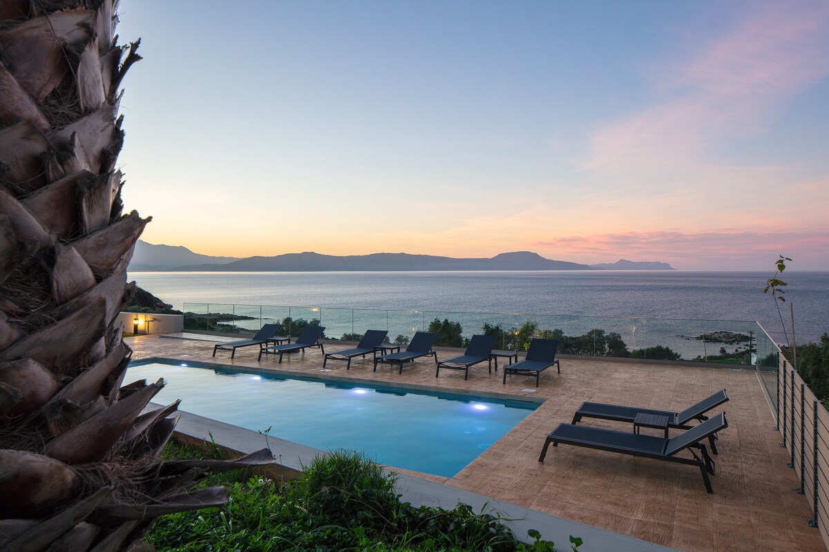 An outdoor area features a serene swimming pool surrounded by lounge chairs. Palm trees line the edge, providing shade. In the background, a tranquil sea and distant mountains can be seen, under a soft evening sky that transitions from blue to warm pink tones.