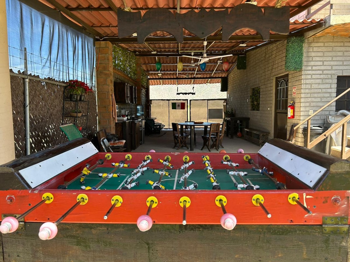 An outdoor recreation area is highlighted by a foosball table, featuring colorful player figures and shiny balls. In the background, a dining table with multiple chairs and a kitchenette can be seen, all sheltered under a roof with terra cotta tiles.