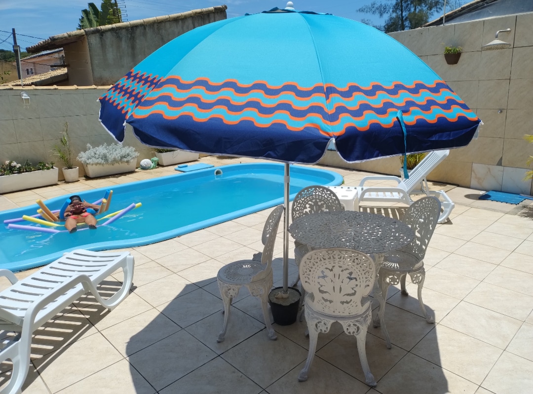 A backyard area features a blue and orange striped umbrella providing shade over a white outdoor table and chairs. A swimming pool is visible in the background, accompanied by colorful pool floats, creating a setting ideal for relaxation.