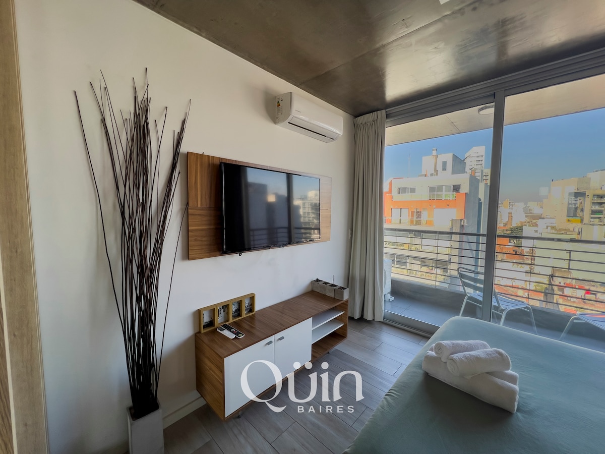 A modern room features a large window with access to a balcony, providing ample natural light. A flat-screen TV is mounted on the wall above a wooden media console with three compartments. Decorative tall branches add a touch of elegance to the contemporary design.