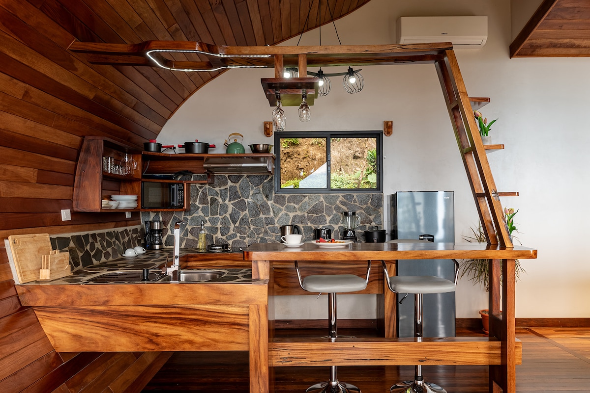 A modern kitchen area showcases wooden cabinetry and stone accents. Stainless steel appliances, including a refrigerator and microwave, are visible. Bar stools are positioned at a curved wooden counter, accompanied by various kitchen tools and utensils, with natural light streaming in through two windows.