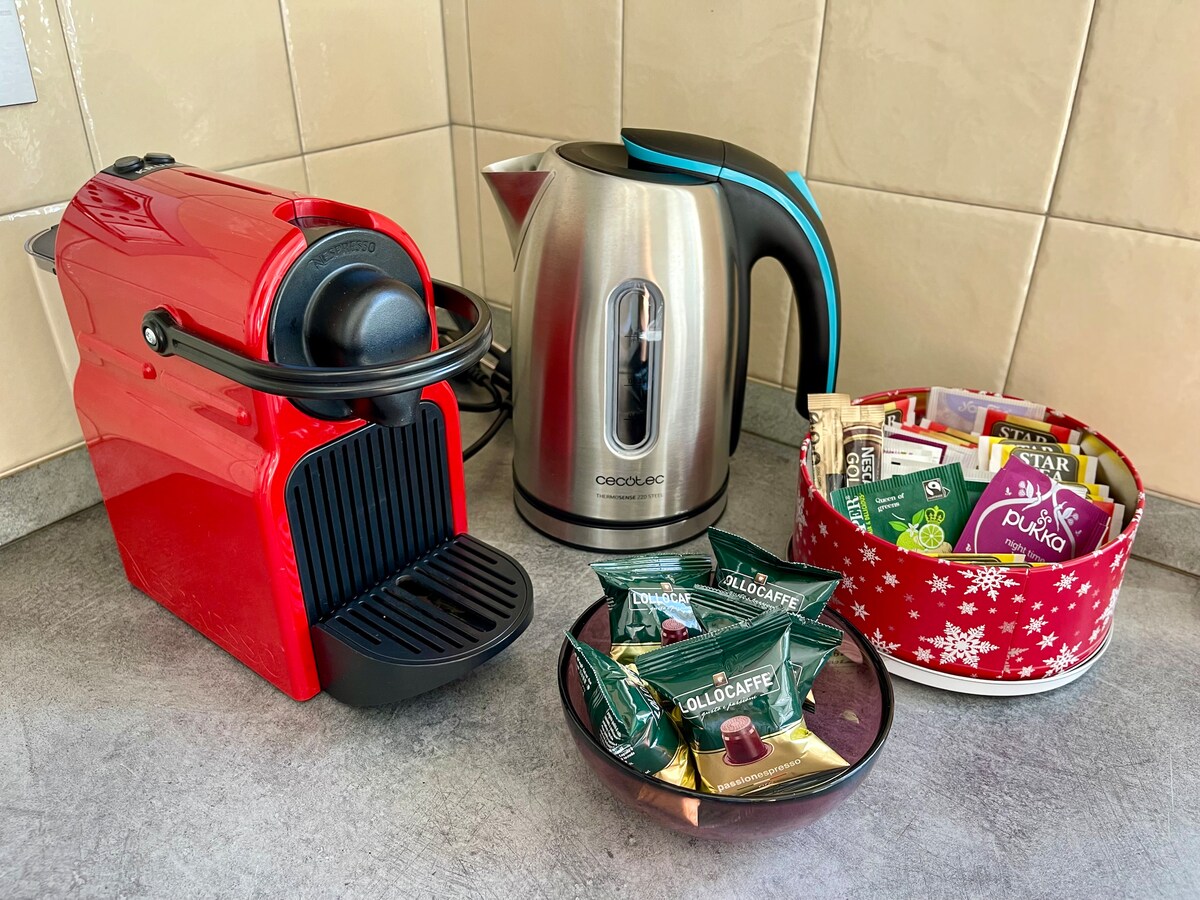 A coffee station is presented with a red espresso machine and a silver electric kettle. Nearby, a bowl filled with assorted coffee capsules is visible, offering a variety of flavors and brands for guests to enjoy.