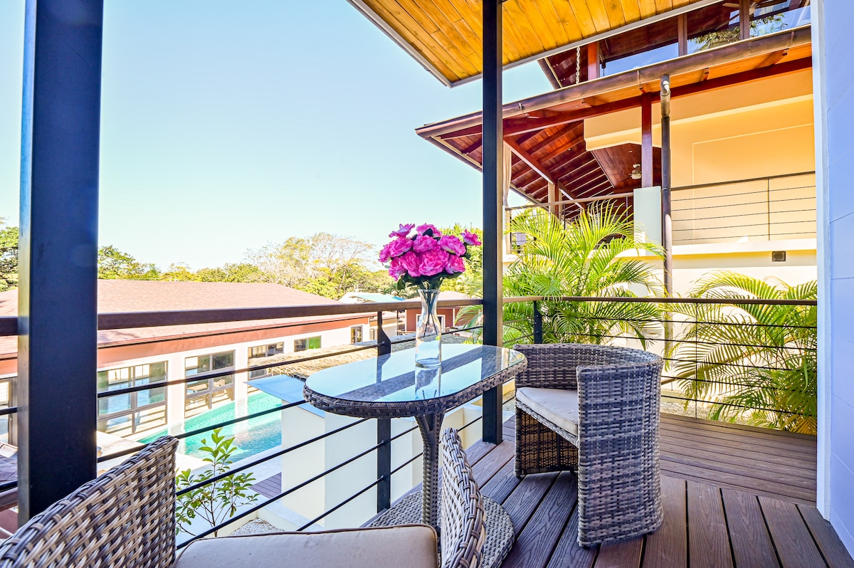 A private balcony features a small glass table accompanied by two woven chairs. A vase with vibrant pink flowers adds a pop of color. In the background, views of the property's infinity pool and lush greenery can be seen, enhancing the outdoor space.