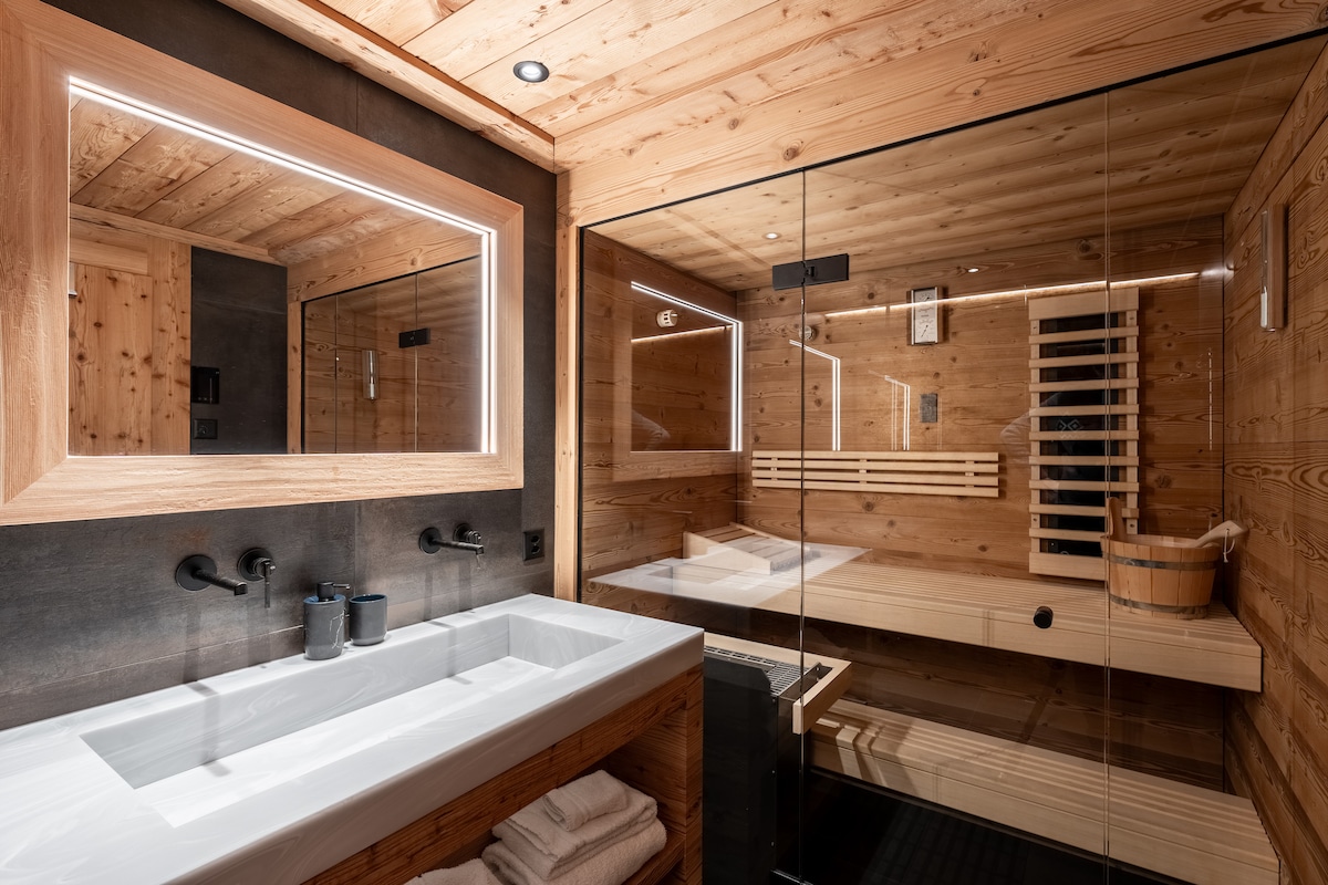 A modern bathroom is complemented by warm wooden accents. A spacious sink area is highlighted with a large mirror, while a sleek sauna with wooden seating is visible in the background. Soft lighting enhances the serene environment, creating a relaxing retreat.