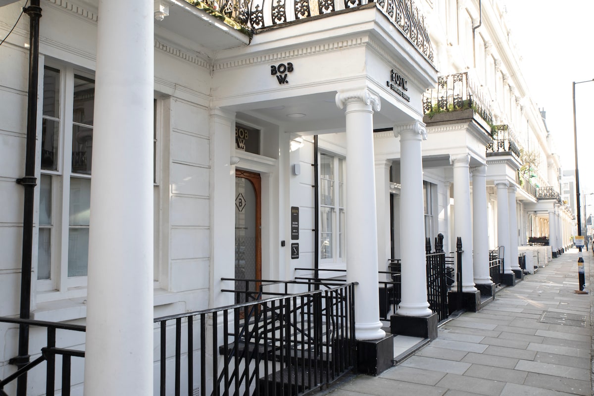 The exterior of the building features classic Georgian architecture with white walls and tall columns. Balconies adorned with planters overlook a paved walkway. A sign marking the entrance is visible, along with decorative railing that frames the area.