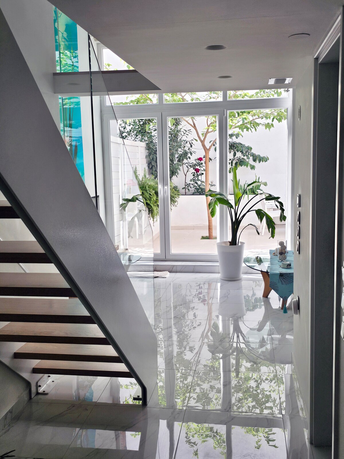 A bright entryway features sleek, polished flooring that reflects light. A staircase with wooden steps leads to the upper level. Large glass doors provide a view of a private garden, with potted plants adding a touch of greenery to the space.