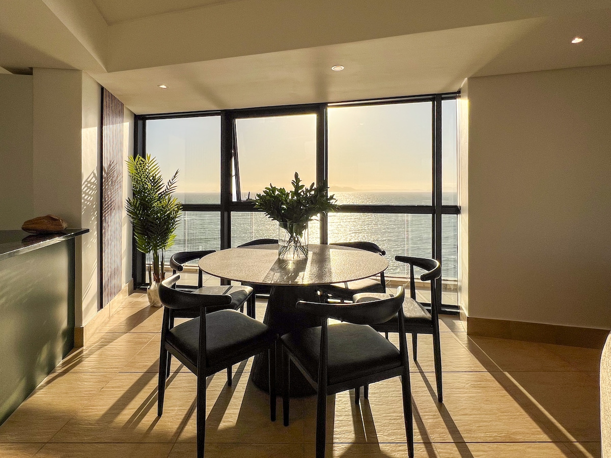 A round dining table is surrounded by four black chairs, set against floor-to-ceiling windows that offer ocean views. Natural light streams in, illuminating the space and casting soft shadows, while a small potted plant adds a touch of greenery.