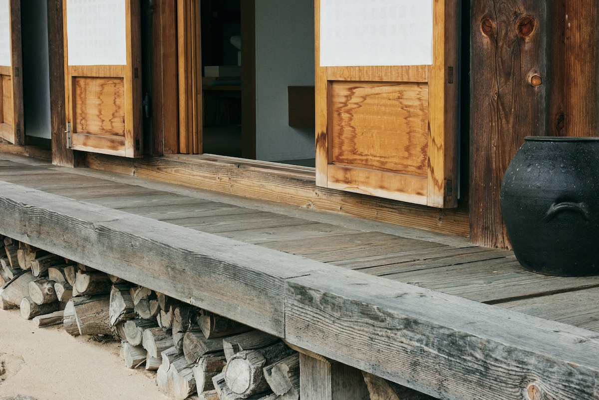 The wooden porch features traditional sliding doors partially open to reveal the interior. A black ceramic pot rests on the floor, and neatly stacked wooden logs are visible underneath, adding to the rustic charm of the space.
