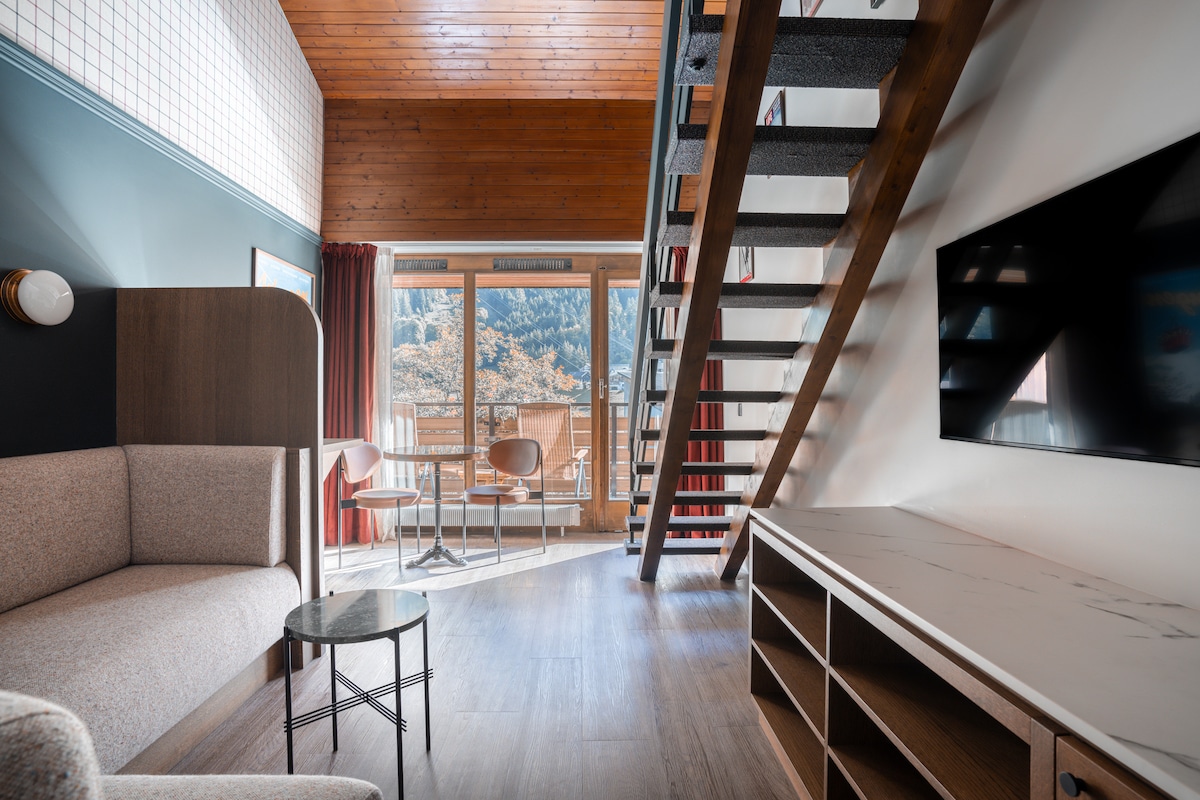 A cozy duplex room features a comfortable seating area with a sofa and a small table. A staircase leads to the upper level, while large glass doors open to a balcony, offering views of the surrounding landscape. A television is mounted on the wall.