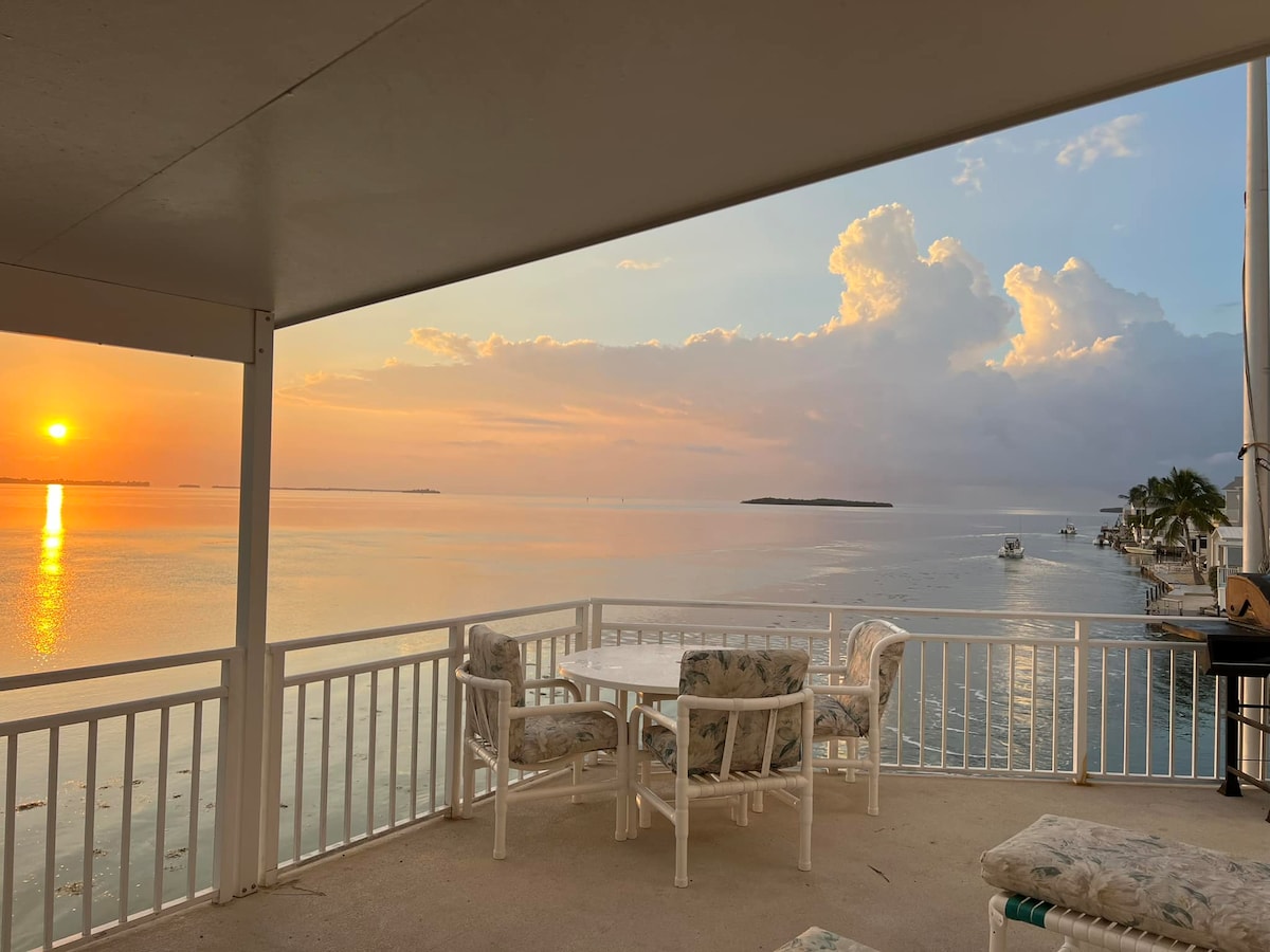 A serene view overlooks the water at sunrise, with soft pastels reflecting on the surface. A white patio table and chairs are positioned under the covered porch, inviting relaxation. The horizon features gentle clouds and distant islands, creating a peaceful atmosphere.