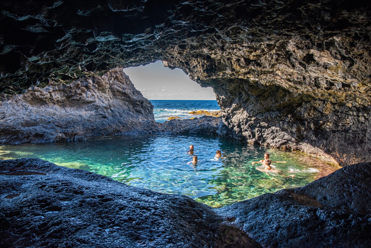A natural rock formation creates an archway leading to a serene pool of clear water. Two people are seen swimming, while rocky shores frame the tranquil scene. The ocean is visible in the background, with gentle waves lapping against the rocks.