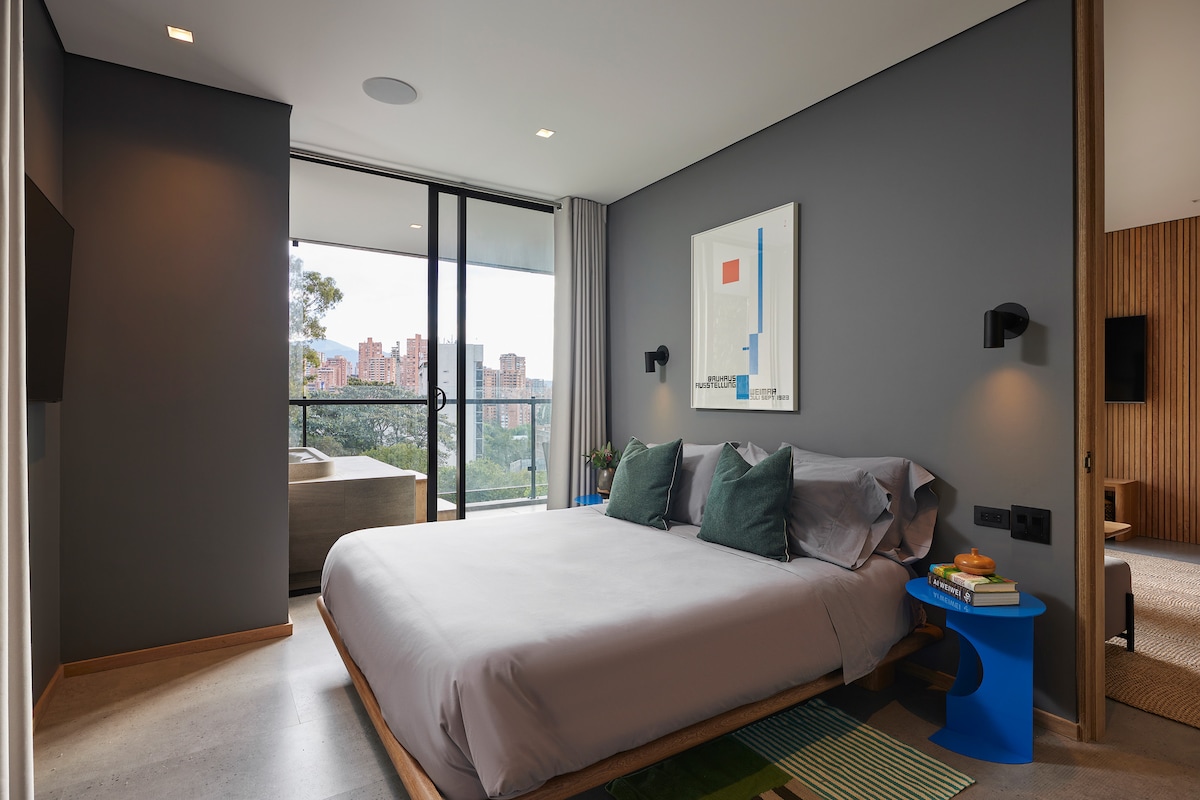 A modern bedroom is showcased, featuring a spacious bed with soft grey linens and decorative pillows in green shades. Large glass doors offer a view of the city, while a stylish piece of artwork hangs on the wall. A small blue table is positioned beside the bed.