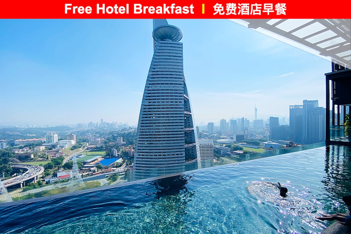 A rooftop swimming pool is shown, with views of the city skyline. The clear blue water reflects the surrounding high-rise buildings. A person is seen swimming, providing a sense of relaxation and spaciousness. The sky is bright, enhancing the openness of the scene.
