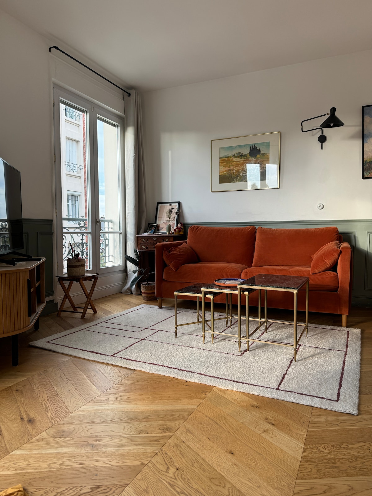 A bright living area is highlighted by a plush orange sofa positioned next to two gold side tables. A wooden cabinet and a television add to the layout, while large windows with sheer curtains allow natural light to fill the space. A textured rug covers the hardwood floor.
