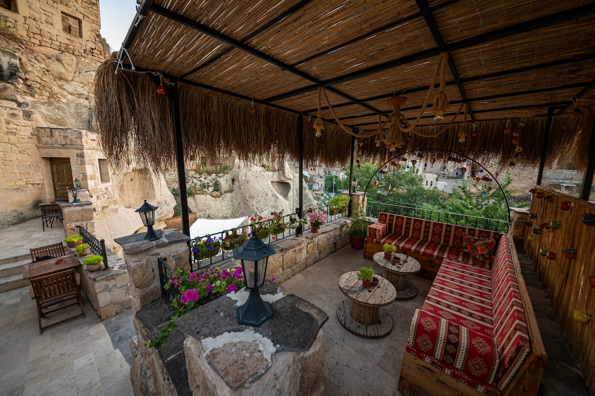 An outdoor seating area is framed by a thatched roof, featuring a comfortable couch adorned with traditional textiles. Decorative lanterns hang overhead, and flower pots are arranged around stone tables, creating a serene space surrounded by natural rock formations.