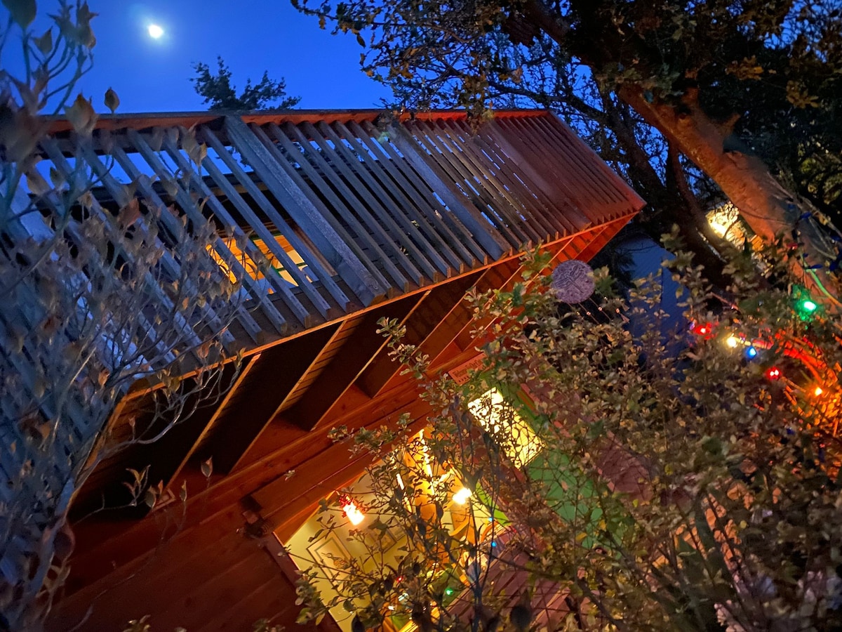 The exterior of the cottage is highlighted at dusk, with a deck featuring wooden railings and festive lights. A moon hangs overhead, and the surrounding greenery adds to the tranquil atmosphere. The cottage's warm interior glow is partially visible through the windows.