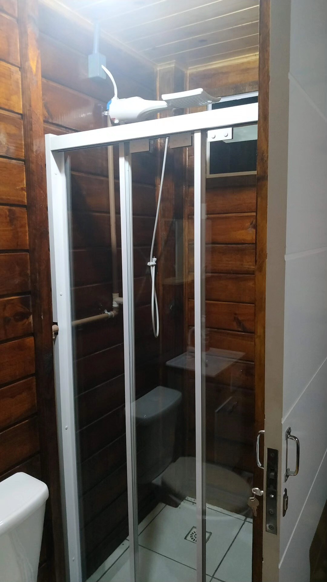 A compact bathroom features a glass-enclosed shower area and a modern toilet. The warm wooden paneling adds a rustic touch, while white tiles provide a clean contrast. Light fixtures above illuminate the space, enhancing visibility.