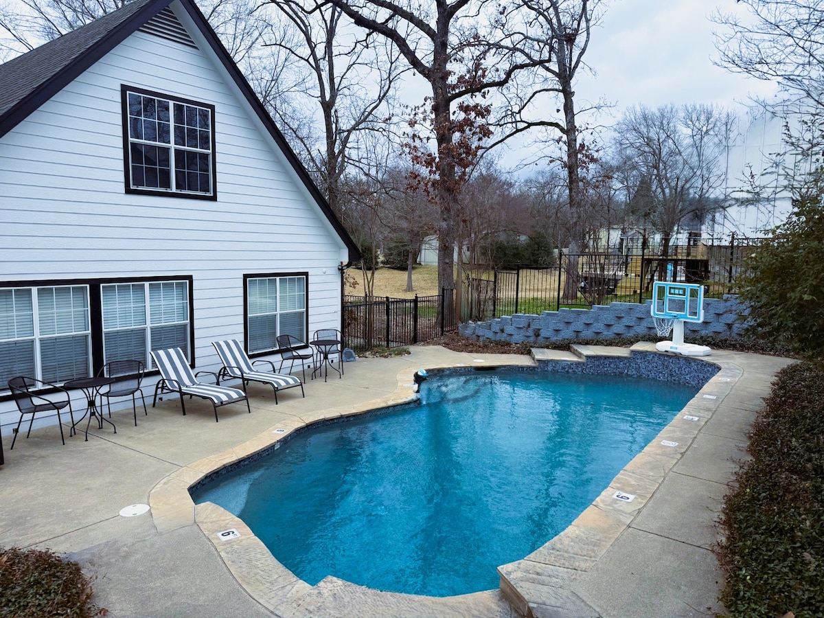 A private pool is set in a fenced backyard, featuring a smooth concrete deck with several lounge chairs positioned alongside. The pool is surrounded by greenery, with a well-maintained lawn visible in the background. A pool house is located adjacent to the pool area.