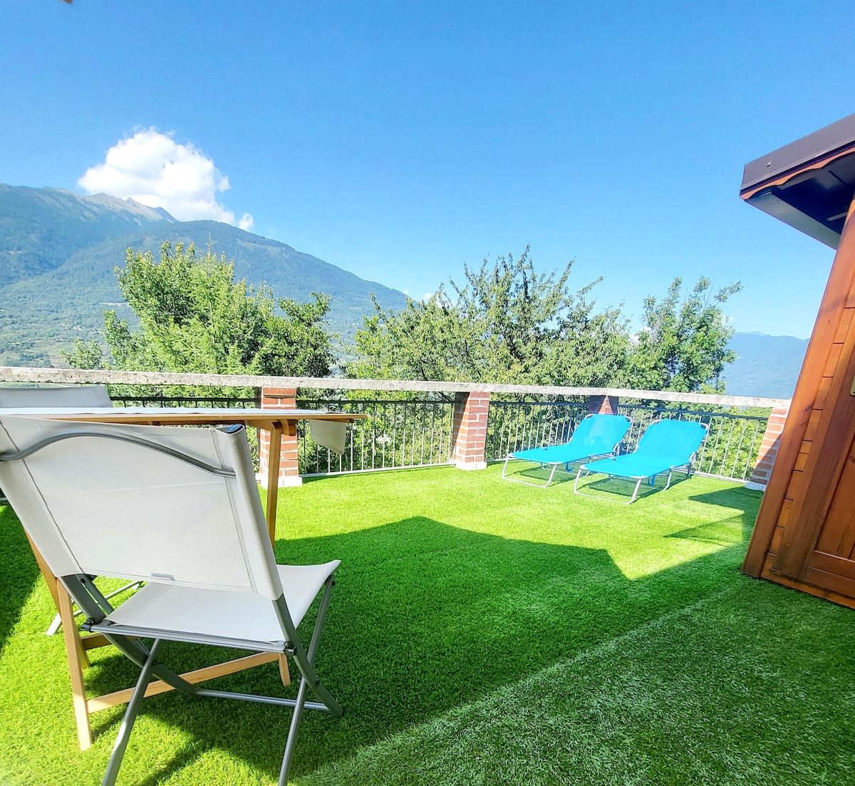 A sunlit outdoor terrace features two blue lounge chairs positioned for relaxation, with a sturdy table nearby. Lush green grass covers the ground, and scenic mountain views are visible in the background under a clear blue sky.