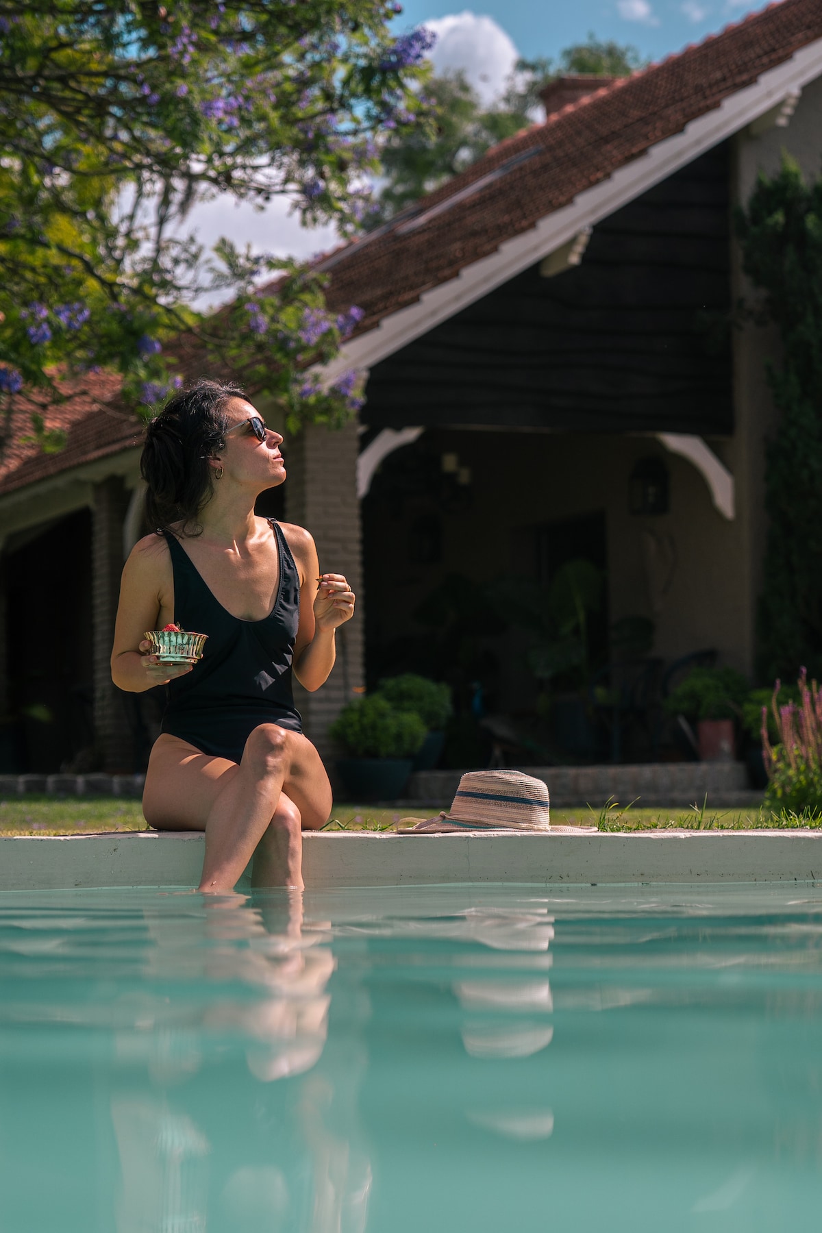 A serene pool area reflects the surrounding greenery, with a clear turquoise water inviting relaxation. A charming house with a rustic wooden facade is visible in the background, framed by blooming plants and trees. A wide-brimmed hat rests nearby, suggesting leisure.