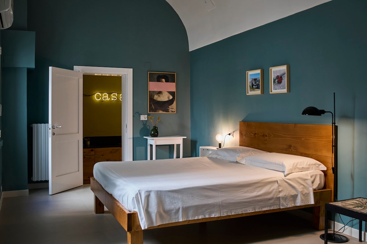 A spacious bedroom features a large wooden bed with crisp white linens, complemented by a bedside lamp and a minimalist nightstand. Soft teal walls are adorned with framed artwork, and a well-lit doorway leads to a welcoming space marked by illuminated signage.