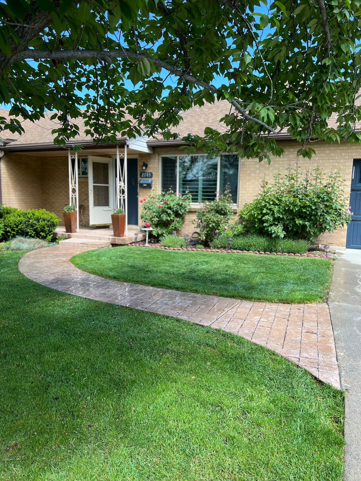 A welcoming entrance is framed by neatly manicured grass and a curved brick pathway. Potted plants add greenery to the front porch area, with a large window revealing the interior beyond. Flowering bushes contribute to the inviting ambiance of the exterior.