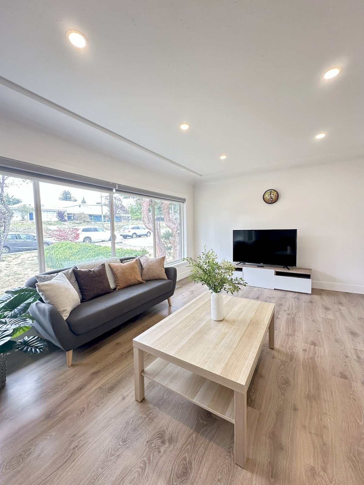 A modern living room features a grey sofa adorned with decorative pillows, positioned near a large panorama window. A light wood table sits centrally in the space. A flat-screen television is mounted on the wall, complemented by minimalist decor and natural light flowing in.