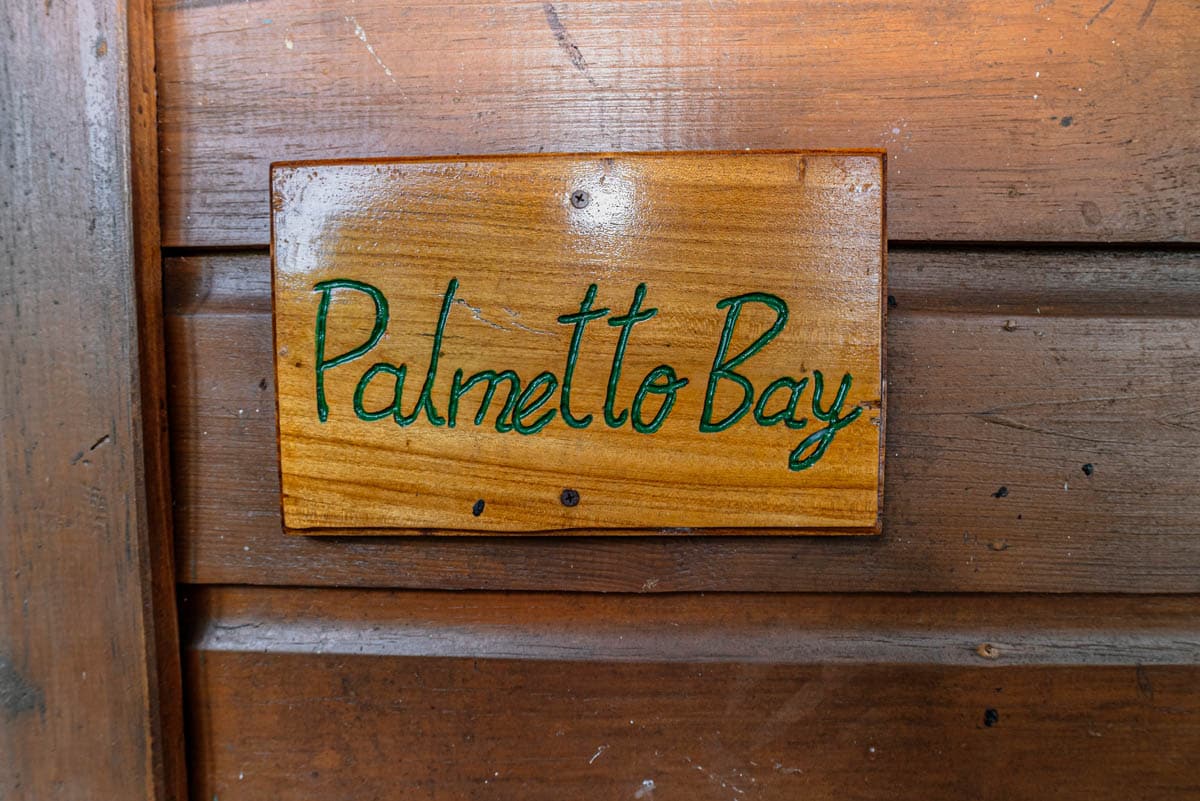 A wooden sign with the inscription 'Palmetto Bay' is displayed on a rustic wooden surface. The lettering is painted in a green hue, contrasting against the natural grain of the wood.