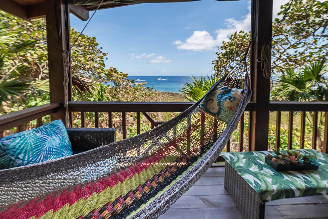 A comfortable hammock hangs between wooden posts on a furnished porch, featuring colorful cushions. The expansive view of the ocean is framed by lush greenery, with small boats visible on the water, under a bright sky with a few clouds.