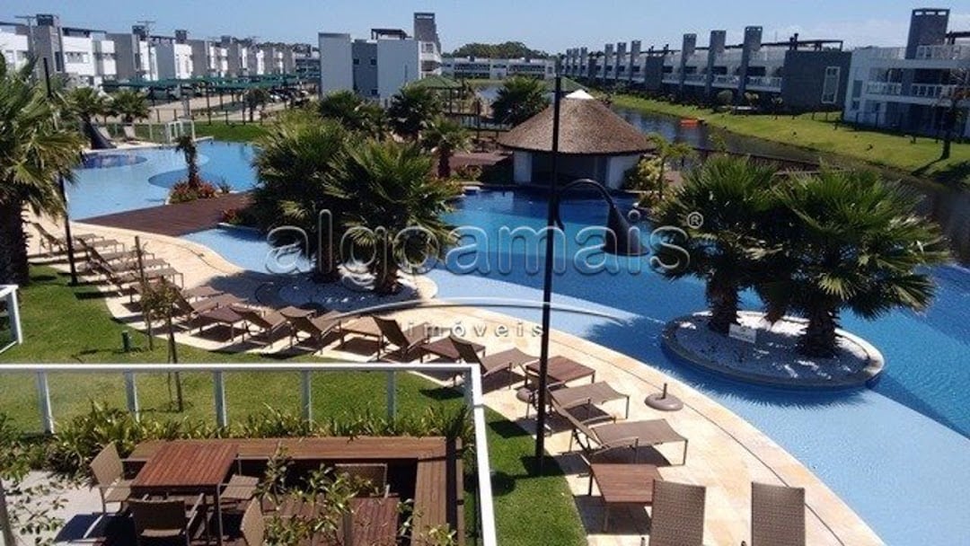 A large outdoor pool area is surrounded by palm trees and lounge chairs, offering ample space for relaxation. Wooden decking complements the serene atmosphere, while a thatched-roof structure provides shade. Multiple residential units are visible in the background, contributing to the community feel.