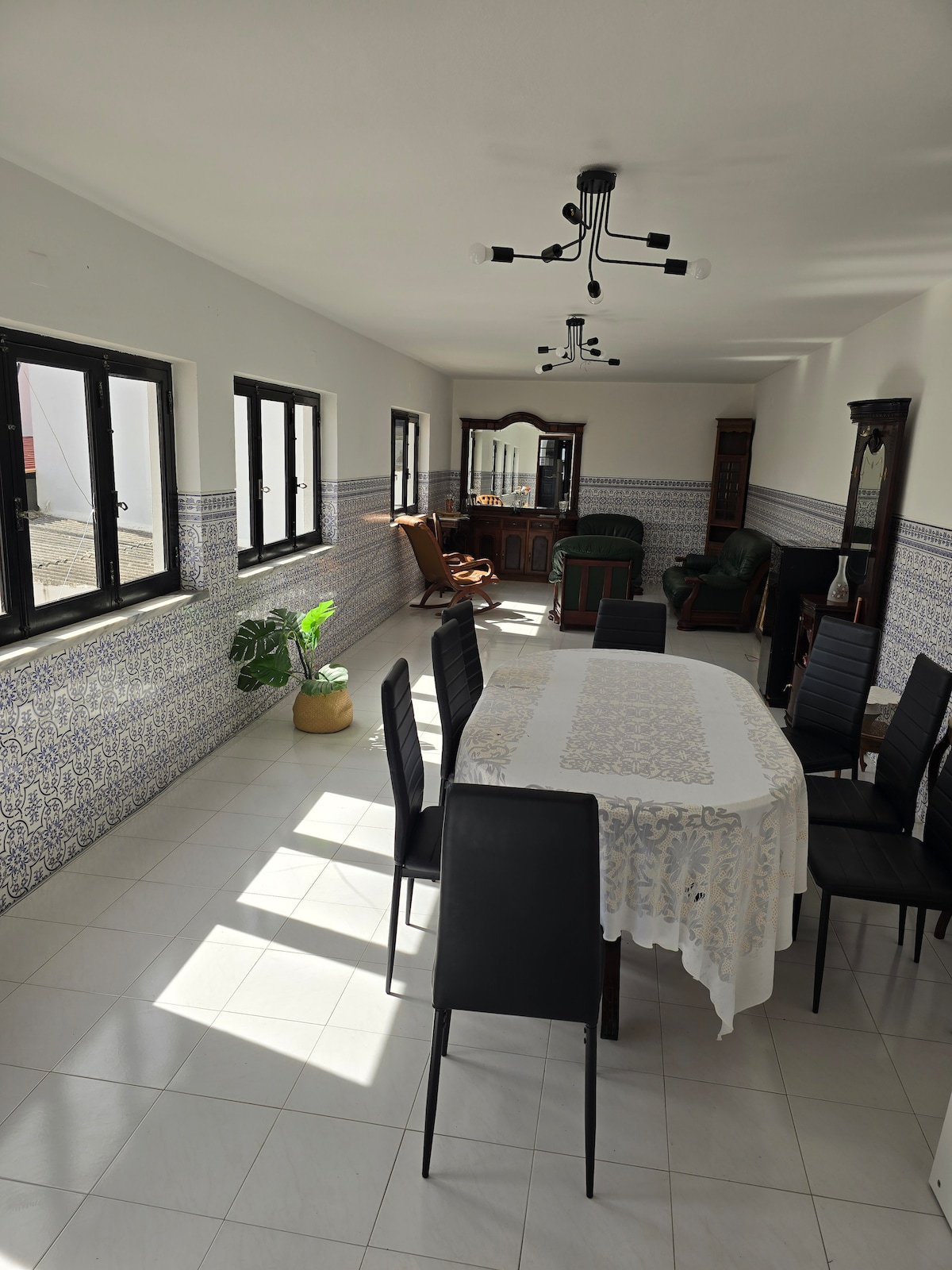A spacious dining area features a large table surrounded by black chairs, with natural light filtering through several windows. Decorative tiles line the walls, and a cozy seating area is visible in the background, showcasing comfortable armchairs and a wooden cabinet.