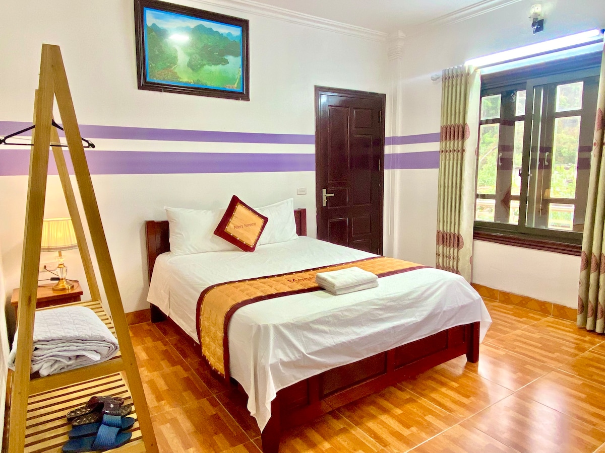 A well-lit guest room features a spacious bed adorned with crisp white linens and a decorative throw. A large window frames a view of the outdoors, while soft curtains complement the room's calming color scheme. A wooden rack displays towels and personal items.