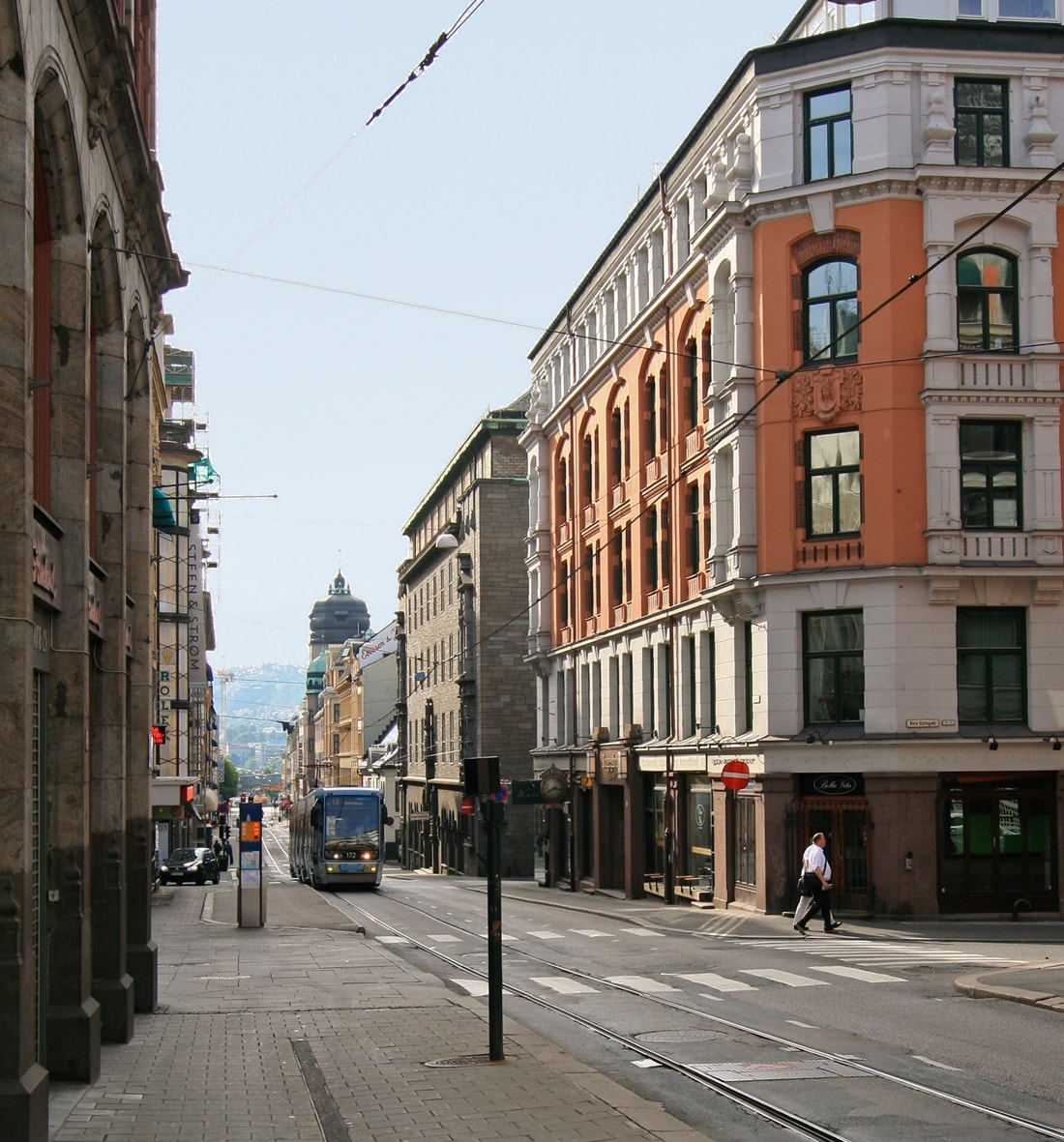 A street view captures a vibrant mix of historic and contemporary architecture along a bustling urban avenue. Tram tracks run down the center, with a tram visible. Pedestrians stroll along the wide sidewalks, complemented by well-maintained buildings featuring various colors and detailed facades.