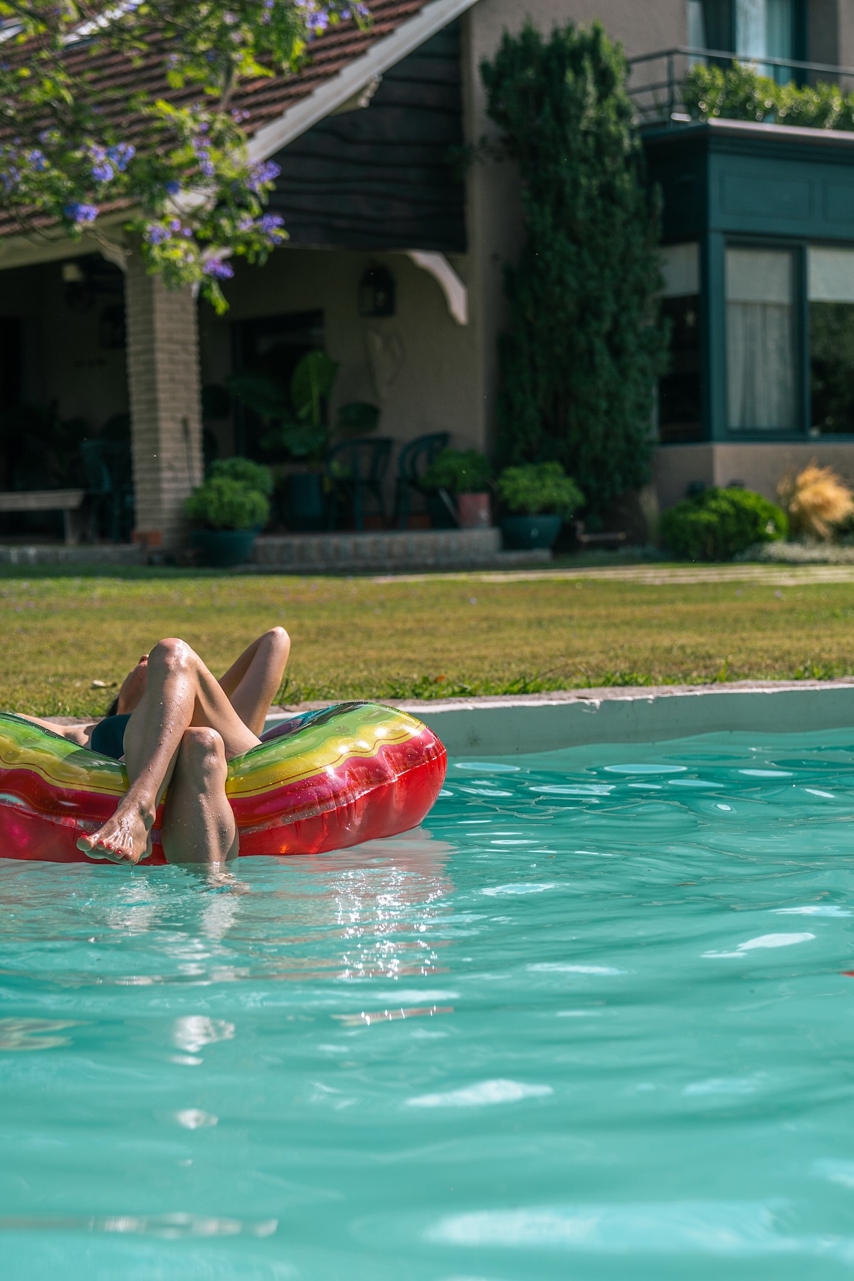 A brightly colored float is gently bobbing on a clear turquoise pool, with a sunbather relaxing on top. The serene scene is complemented by the greenery of a well-maintained yard and the exterior of an inviting house in the background.
