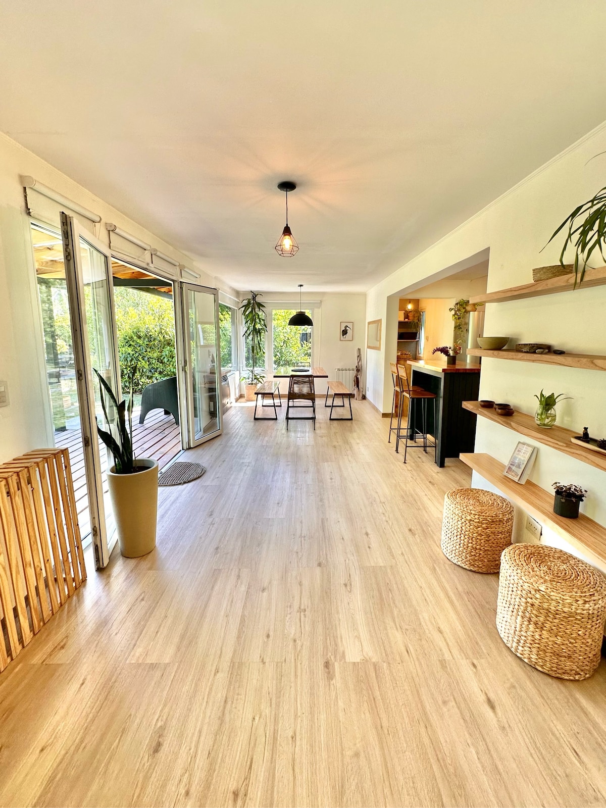 A bright and spacious living area features large glass doors leading to a garden. Natural light fills the space, accentuating the wood flooring. A dining table is set for four, with additional seating nearby. Decorative plants add a touch of greenery throughout.