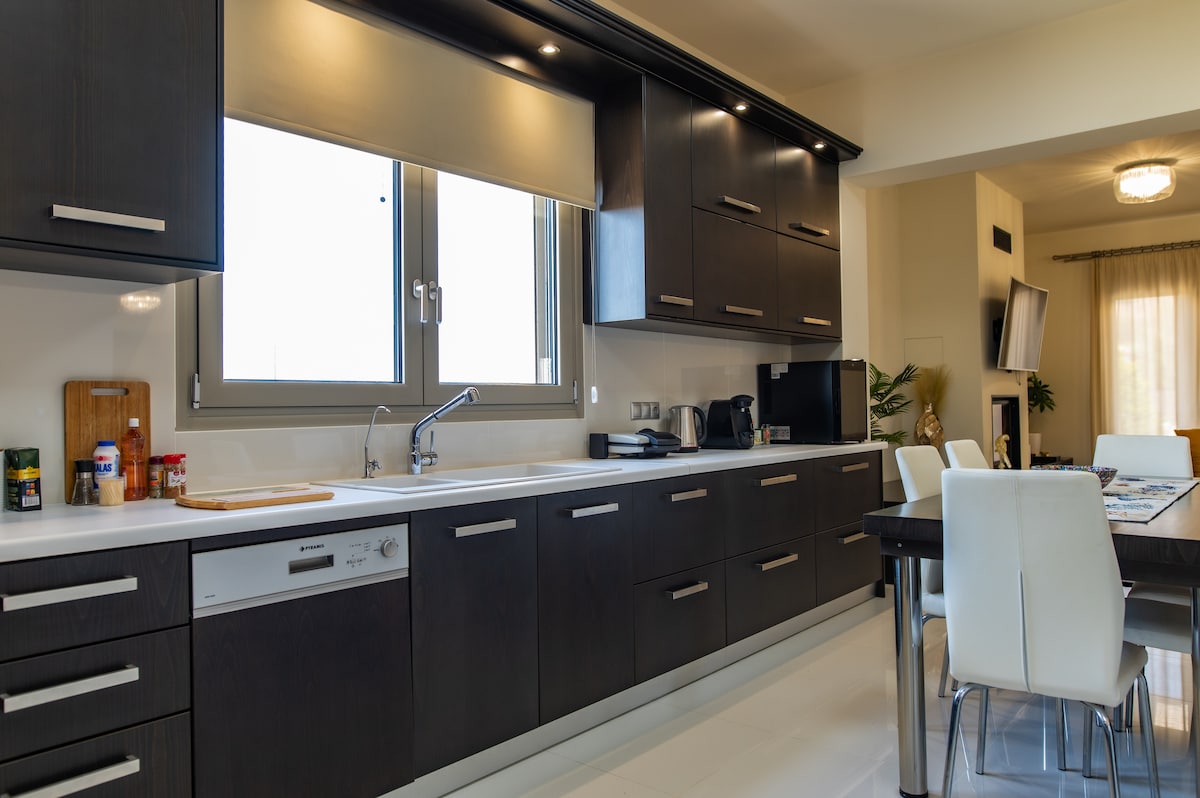 A modern kitchen with dark wood cabinetry and a sleek countertop is designed for comfort and functionality. Equipped with essential appliances, including a dishwasher and kettle, this space is complemented by a dining table surrounded by white chairs, creating an inviting meal preparation area.