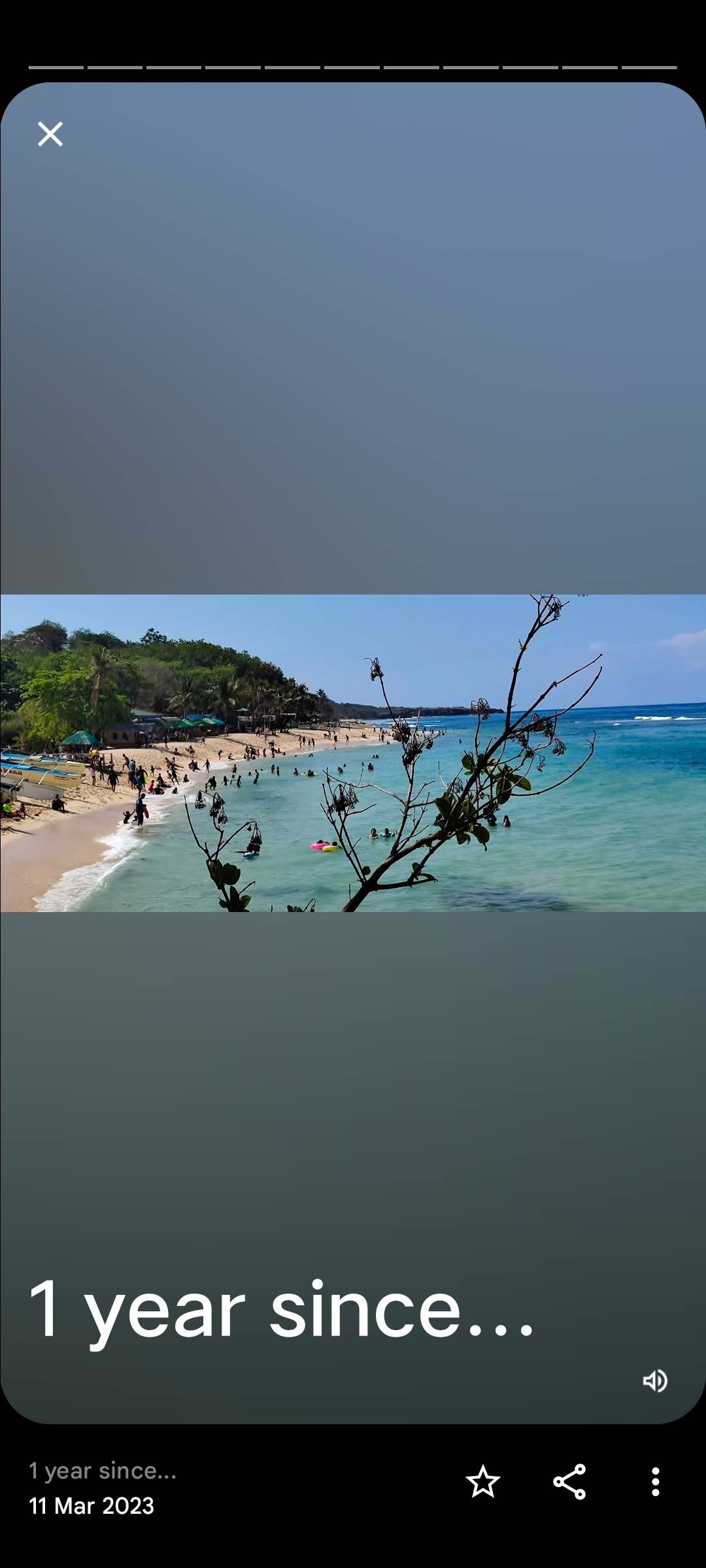 A sandy beach is visible, with visitors enjoying the sun and water along the shoreline. Gentle waves lap at the beach, while hills lined with greenery rise in the background. A few scattered trees frame the scene, contributing to the coastal atmosphere.