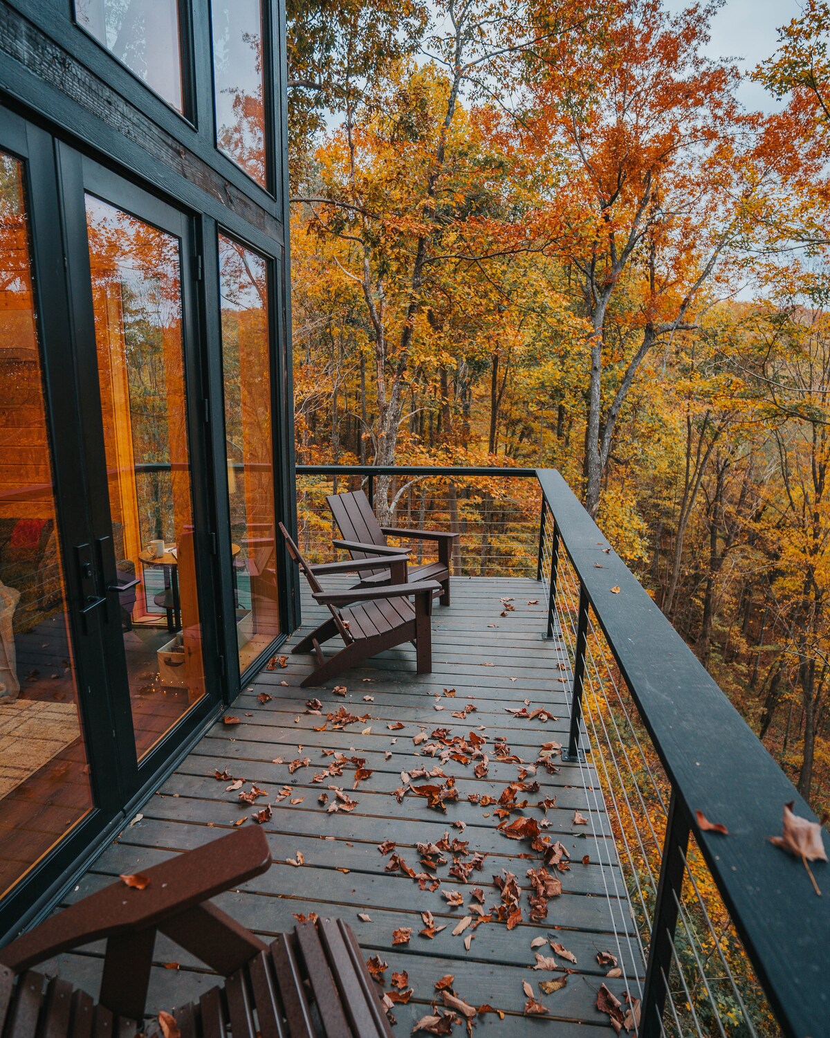 An outdoor balcony is visible with two wooden chairs positioned along the railing, surrounded by autumn foliage. The floor is scattered with orange and brown leaves. Large glass doors reflect the colorful trees outside, enhancing the connection to nature.