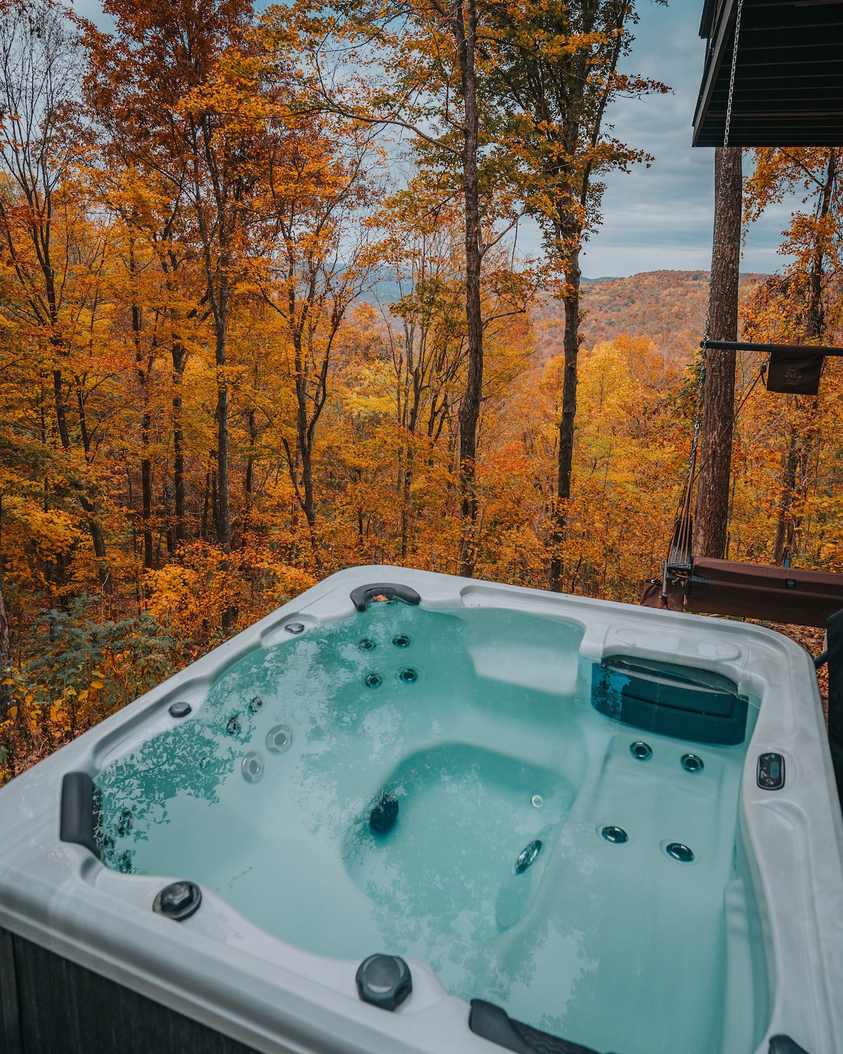 A hot tub is positioned on a deck surrounded by trees with vibrant autumn foliage. The clear water reflects the natural beauty of the landscape, offering a serene view of the mountains in the distance.