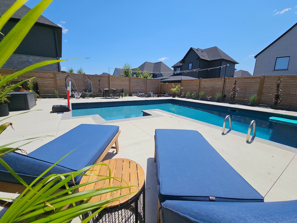 A spacious private backyard features a large swimming pool surrounded by lounge chairs and a small table. Lush greenery adds natural elements to the space, while a fence offers privacy. Bright sunshine enhances the inviting atmosphere, perfect for relaxation.