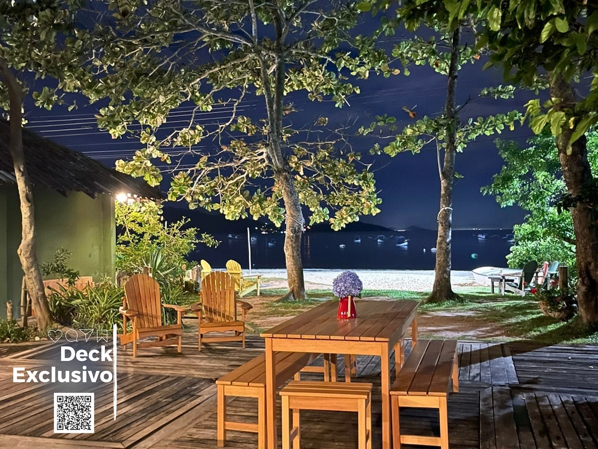 A wooden deck featuring a large table surrounded by outdoor chairs is illuminated by soft lighting. Trees provide shade, and the beach is visible in the distance, enhancing the serene atmosphere of the space during nighttime.