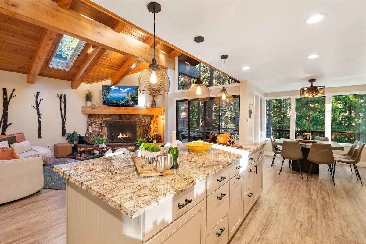 The open-concept living space features a warm kitchen island topped with granite, complemented by pendant lighting and natural wood accents. A cozy seating area is seen beside a stone fireplace, while large windows showcase views of the surrounding forest. A dining area with modern chairs is visible beyond.