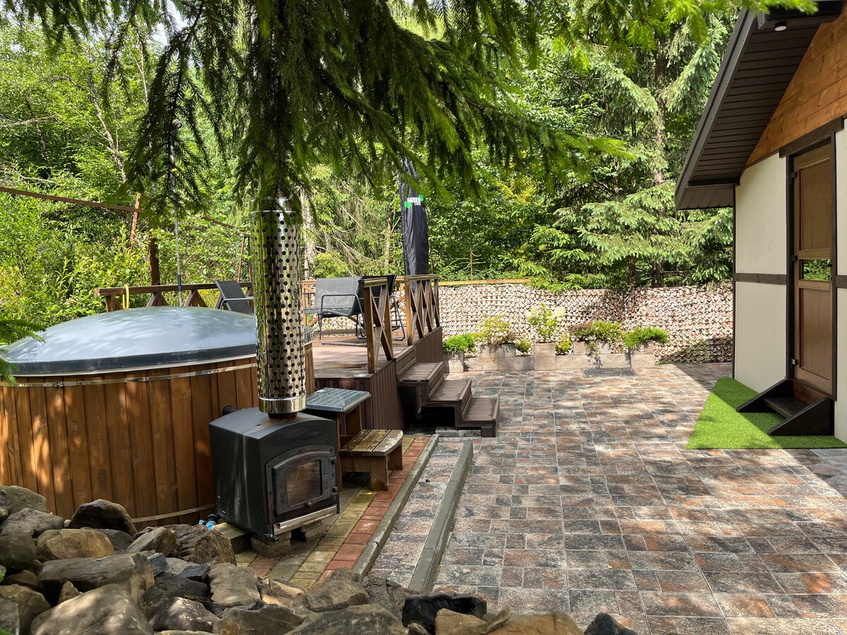 A wooden deck is complemented by a hot tub and a wood stove, surrounded by lush greenery. A stone pathway leads through the outdoor space, with potted plants adding subtle detail. A patio area can be seen in the background, enhancing the connection with nature.