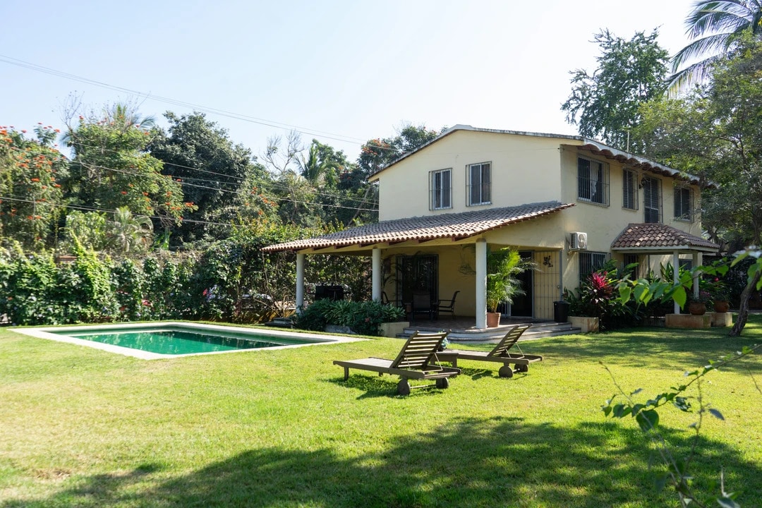 A spacious house is set amidst a lush green garden. A tranquil swimming pool is visible in front, flanked by two lounge chairs. The shaded porch features a tiled roof, providing a comfortable space to relax and enjoy the garden views.